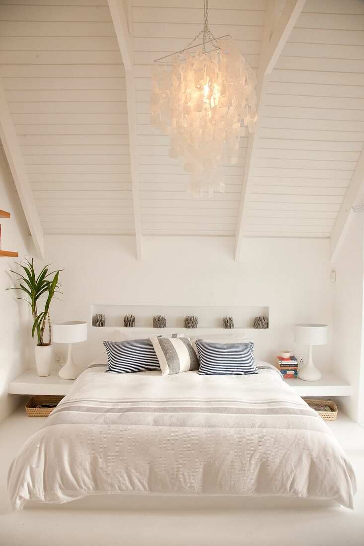 Gemütliches, weisses Doppelbett unter weiss gestrichenem Holzdach; an der Decke eine leuchtende Hängelampe aus Perlmutt