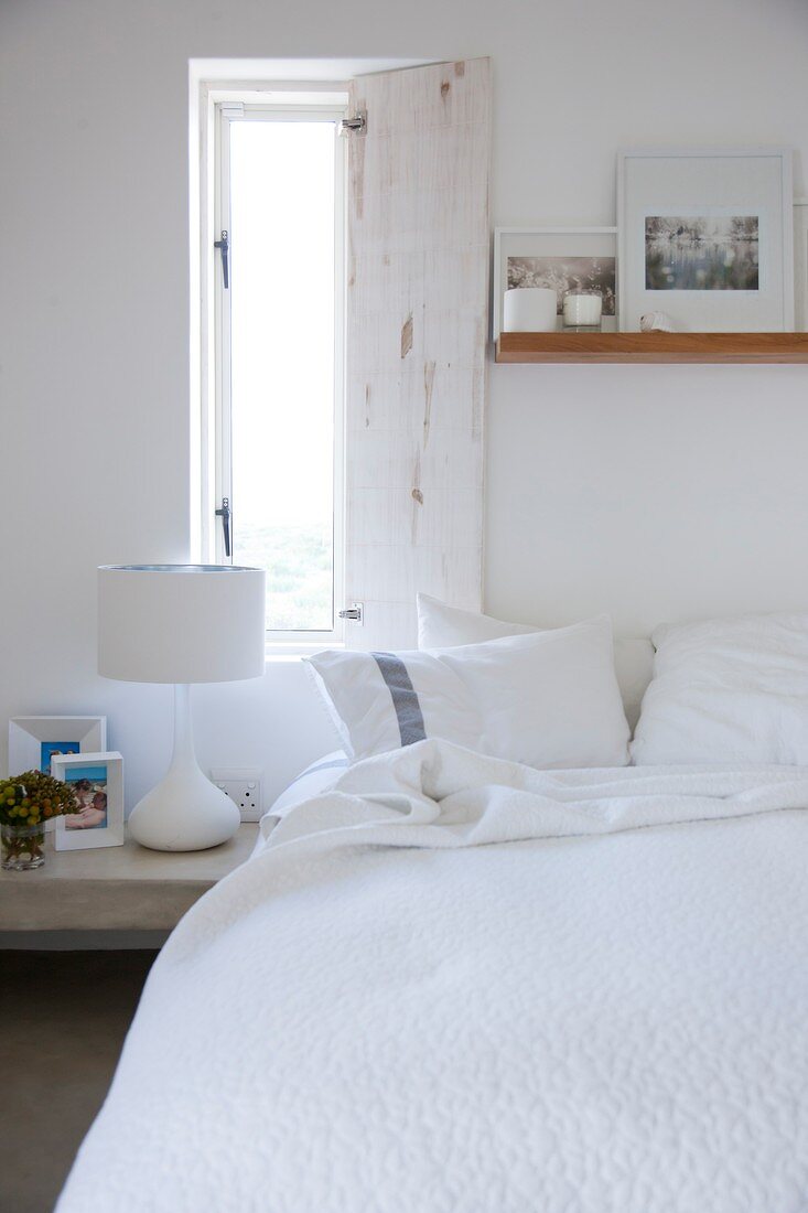 Open window shutter above bed with white bedspread and photos leaning against wall on floating wooden shelf