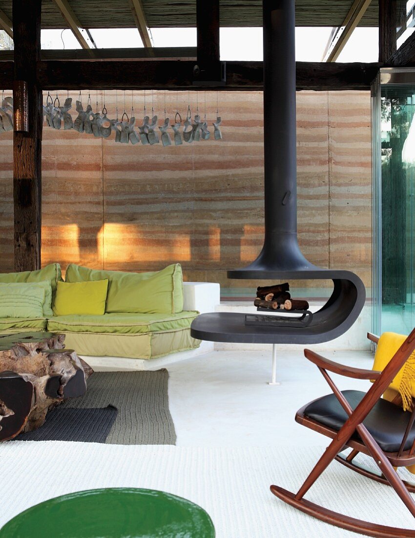 Floating wood-burning stove in modern living room with spring green couch and rustic, root-wood coffee table in front of rammed earth wall