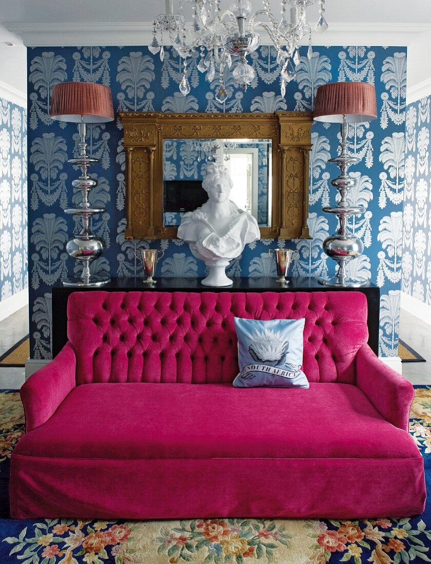 Ausladende, pinkfarbene Couch in luxuriösem Raum mit großgemusterter Wandtapete, Kristallleuchter und Seidenteppich mit großem Blumenmuster