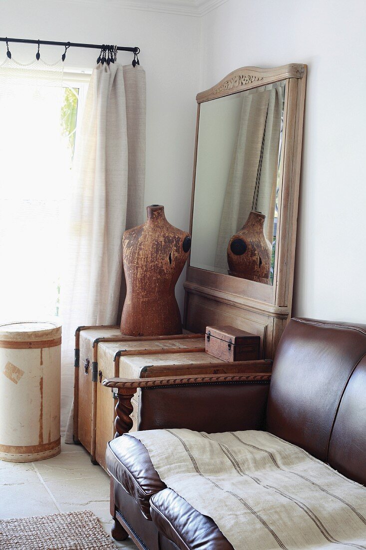 Traditionelles, braunes Ledersofa neben Holztruhe und Schneiderpuppe vor gerahmtem Spiegel in Zimmerecke neben dem Fenster