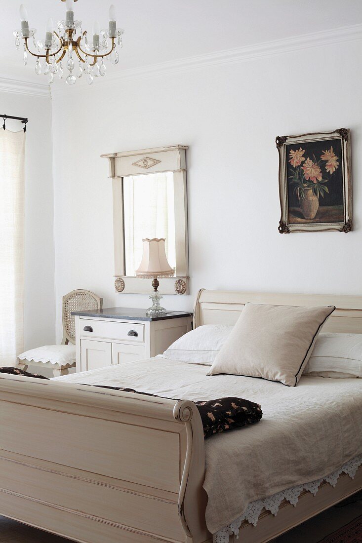 Antikes Schlittenbett in hellem Farbton neben schlichtem Nachtkästchen und gerahmtem Spiegel an Wand