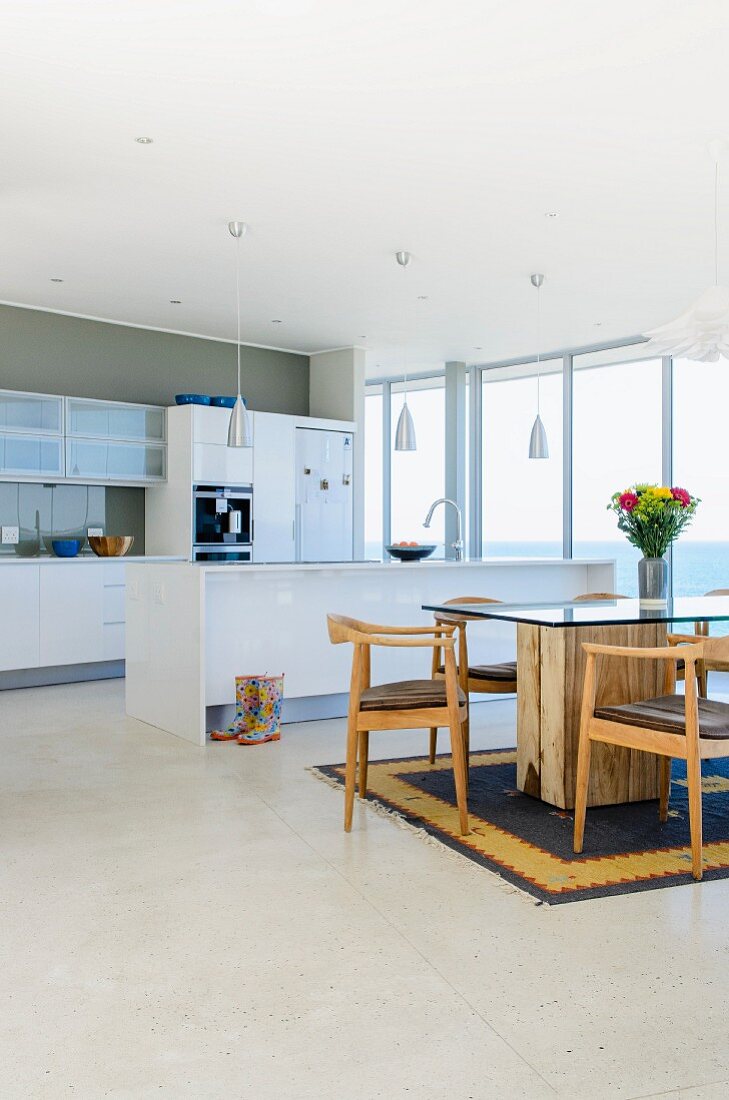 Open-plan interior with island counter in modern, white kitchen; glass dining table with massive wooden base and wooden chairs on Berber rug in foreground