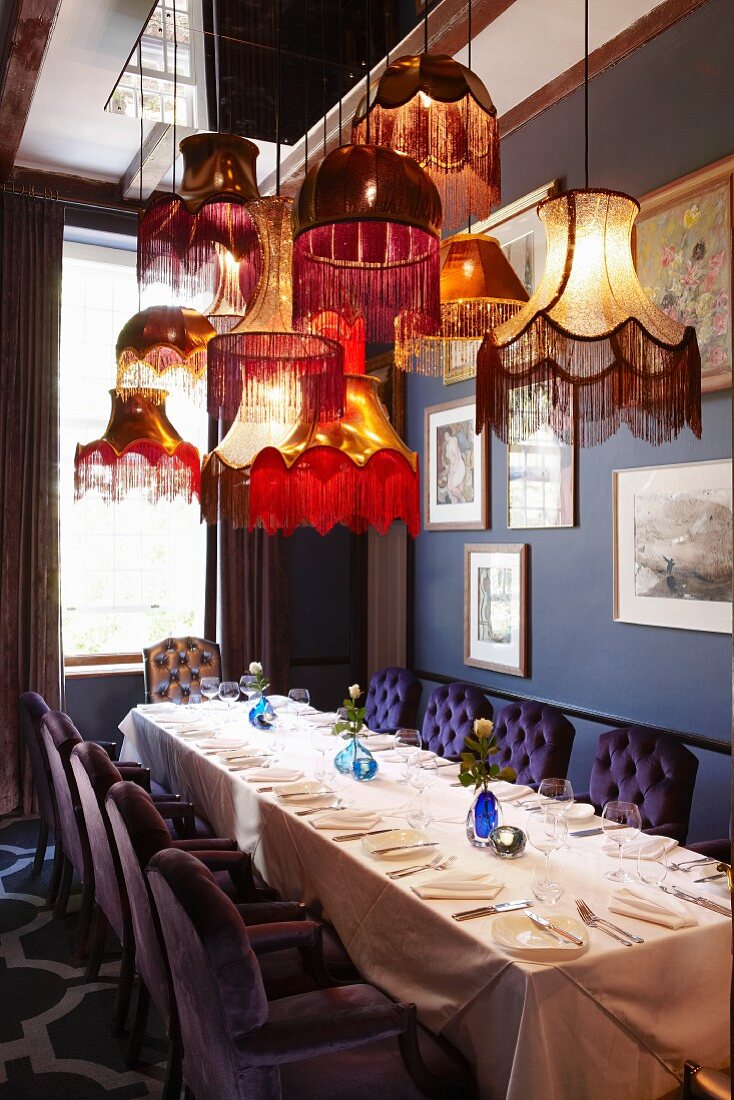 Long, set dining table below collection of retro lampshades; chairs with purple quilted backs harmonising with blue walls