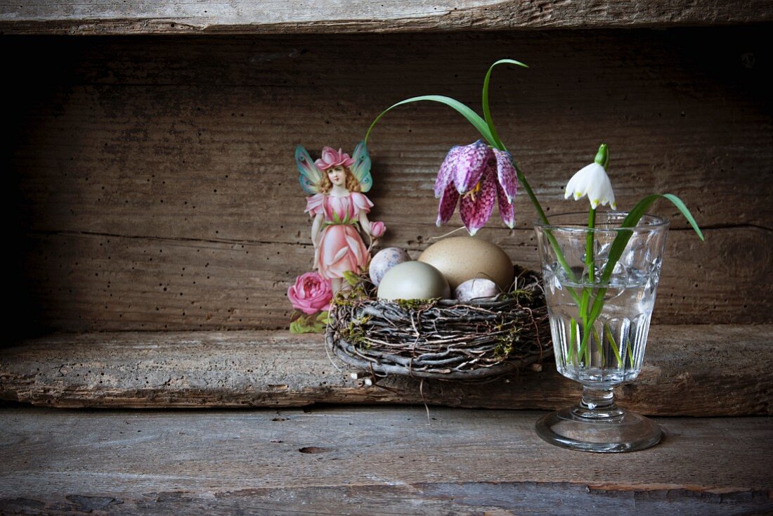 Stillleben mit Nest und verschiedenen Eiern, Schachbrettblume und Schneeglöckchen im Glas und figürliches Klebebild in altem Holzkasten