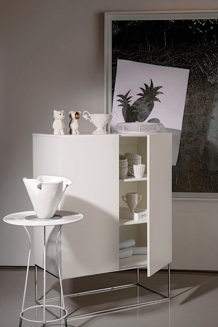 Designerschrank mit weißem Geschirr und Dekogegenständen sowie ein Beistelltischchen mit Vase vor einem zeitgenössischen Wandbild