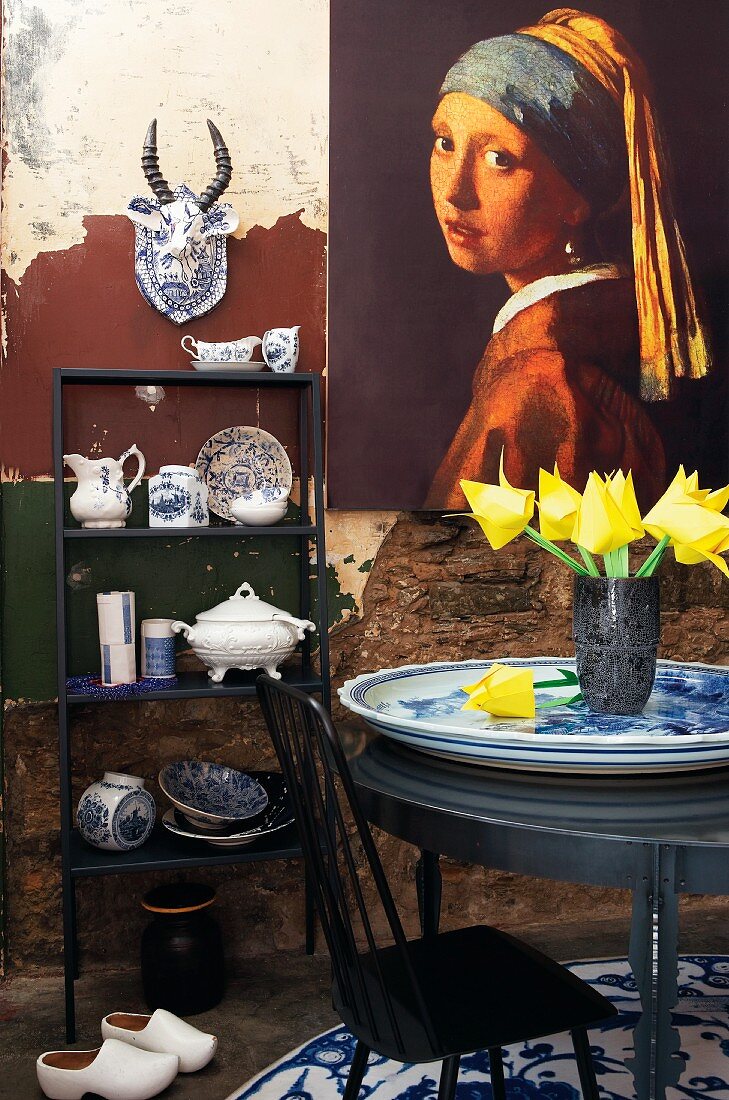 Schränkchen mit holländischem Porzellangeschirr und Dekogegenständen, Wandgemälde und Tisch mit Tulpenstrauss