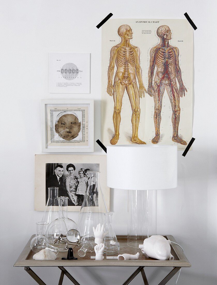 Various drawings and photos on wall above small table holding lamp, laboratory glassware and china objects