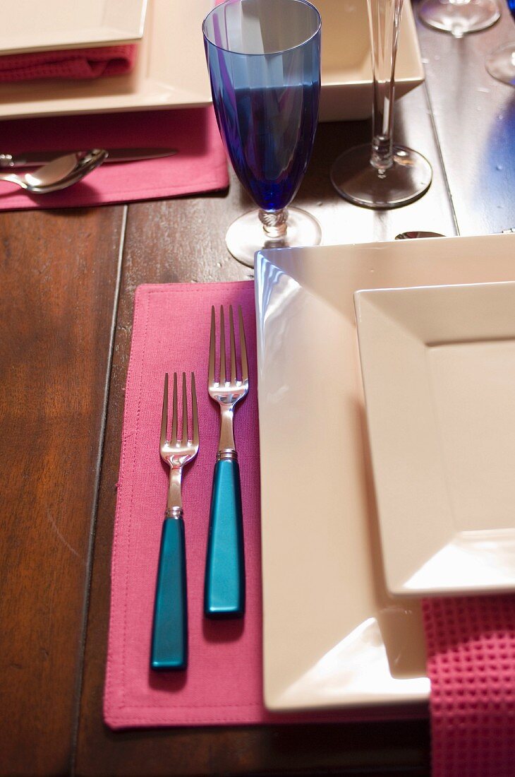 Gedeck mit viereckigen Tellern und Besteck mit blauem Griff auf rosa Tischset