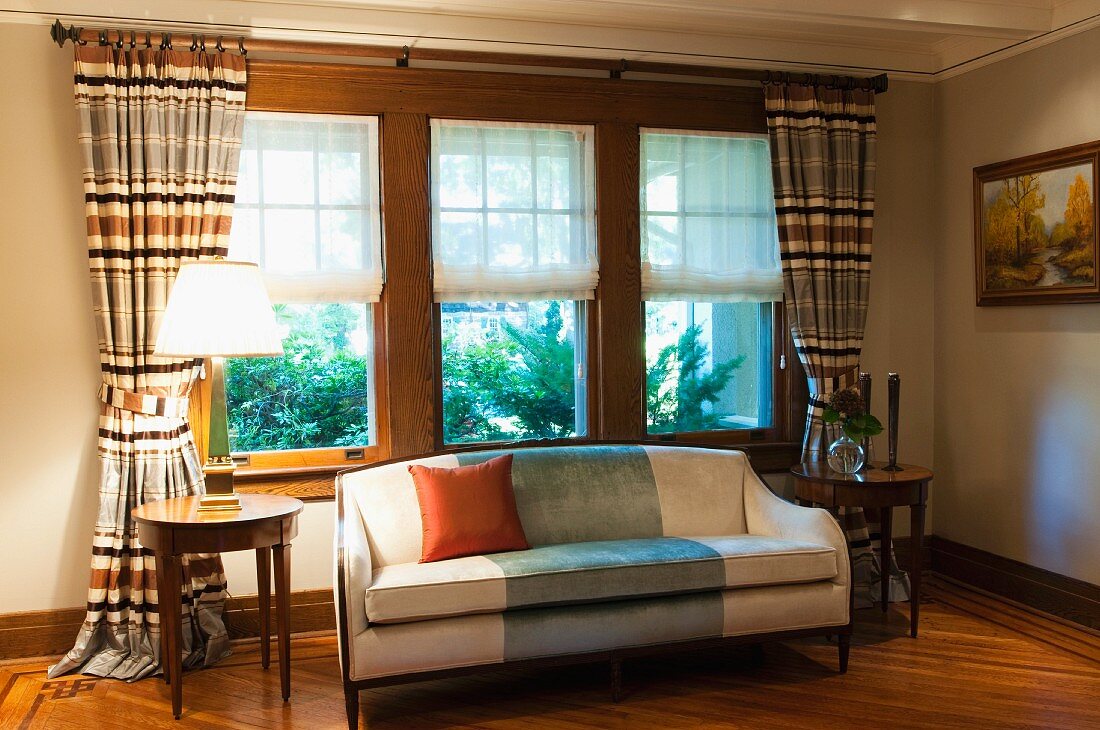 Helles Sofa und runde Beistelltische vor Fenster mit halboffenen Rollos und gerafften Vorhängen in traditionellem Wohnzimmer