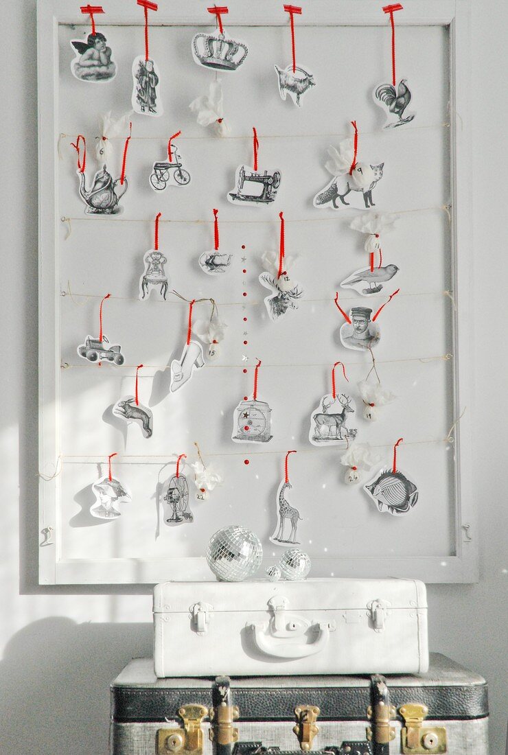 Selbstgebastelter Adventskalender mit verschiedenen Motiven aus Papier und rotem Band auf Wandbord, davor ein Kofferstapel