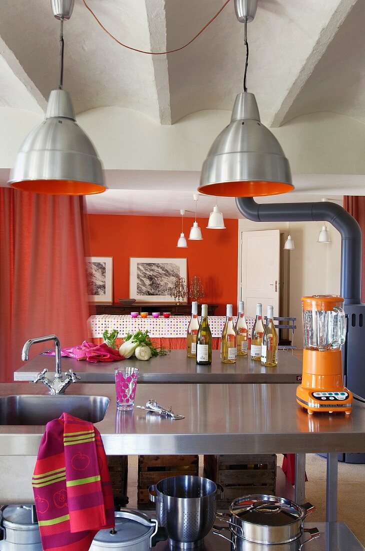 Lampen im Industrielook über Edelstahlspülentisch; orangerot getönte Wand und Vorhang als Raumteiler im Hintergrund