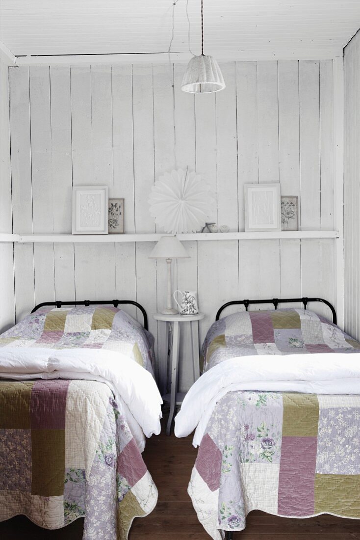 Metal beds with patchwork quilts in front of a wood wall painted white with a shelf