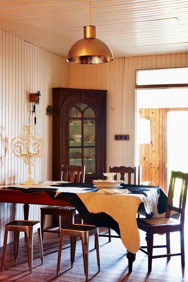 Antique chairs and metal stools around a dining table covered with an cowhide rug; china hutch in front of a wood paneled wall