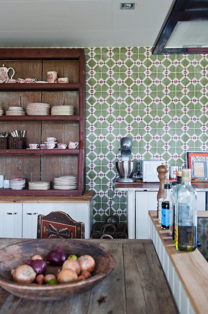 Wooden dish of onions on rustic table next to oil bottles on island counter in front of shelving against wall with patterned tiles