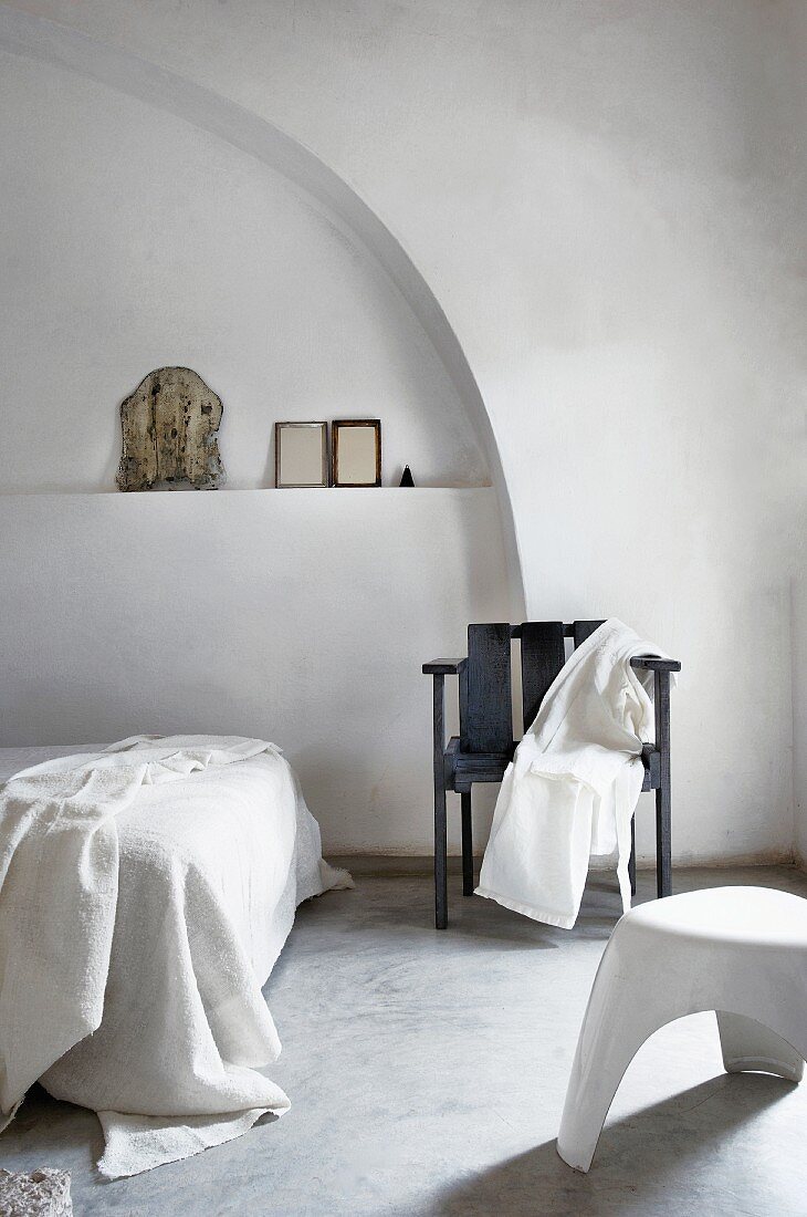 Schwarzer Armlehnenstuhl vor schlicht dekorierter Wandnische und moderner weißer Kunststoffhocker in puristischem Schlafraum