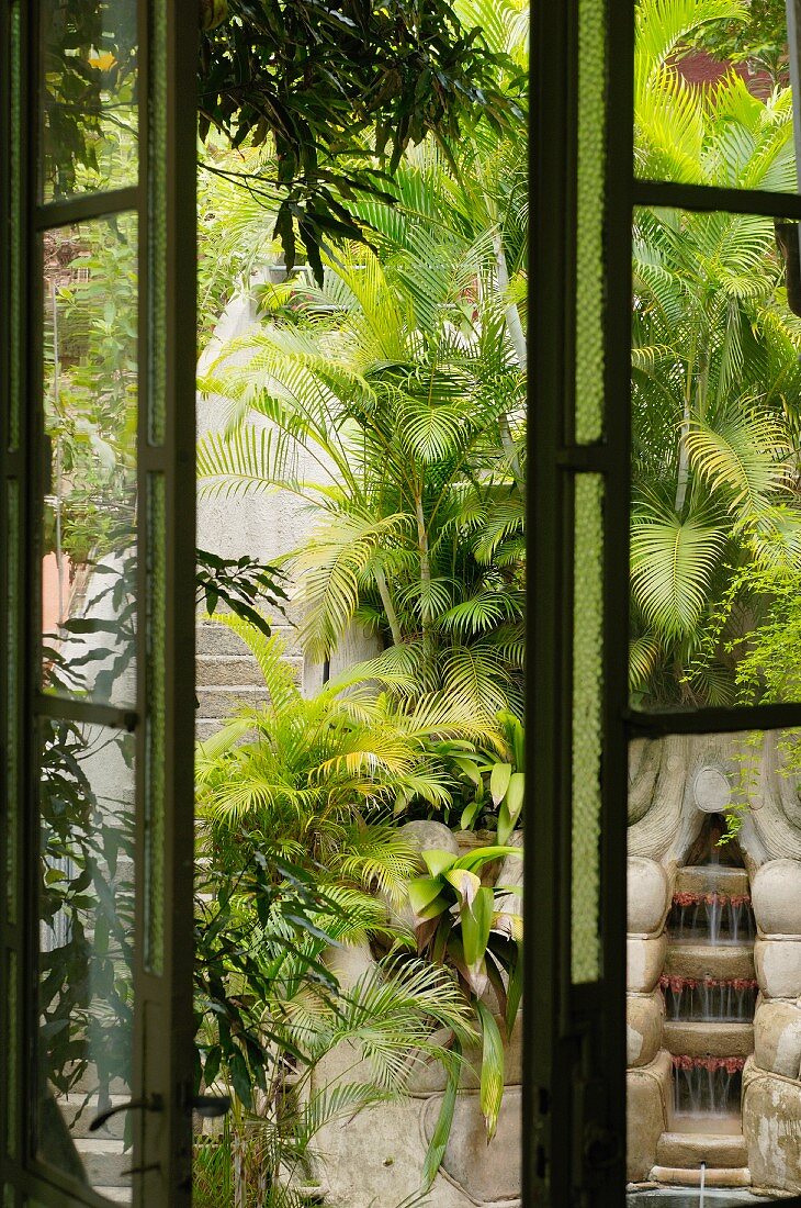 View through half-open window of stone steps and cascade fountain surrounded by palm trees