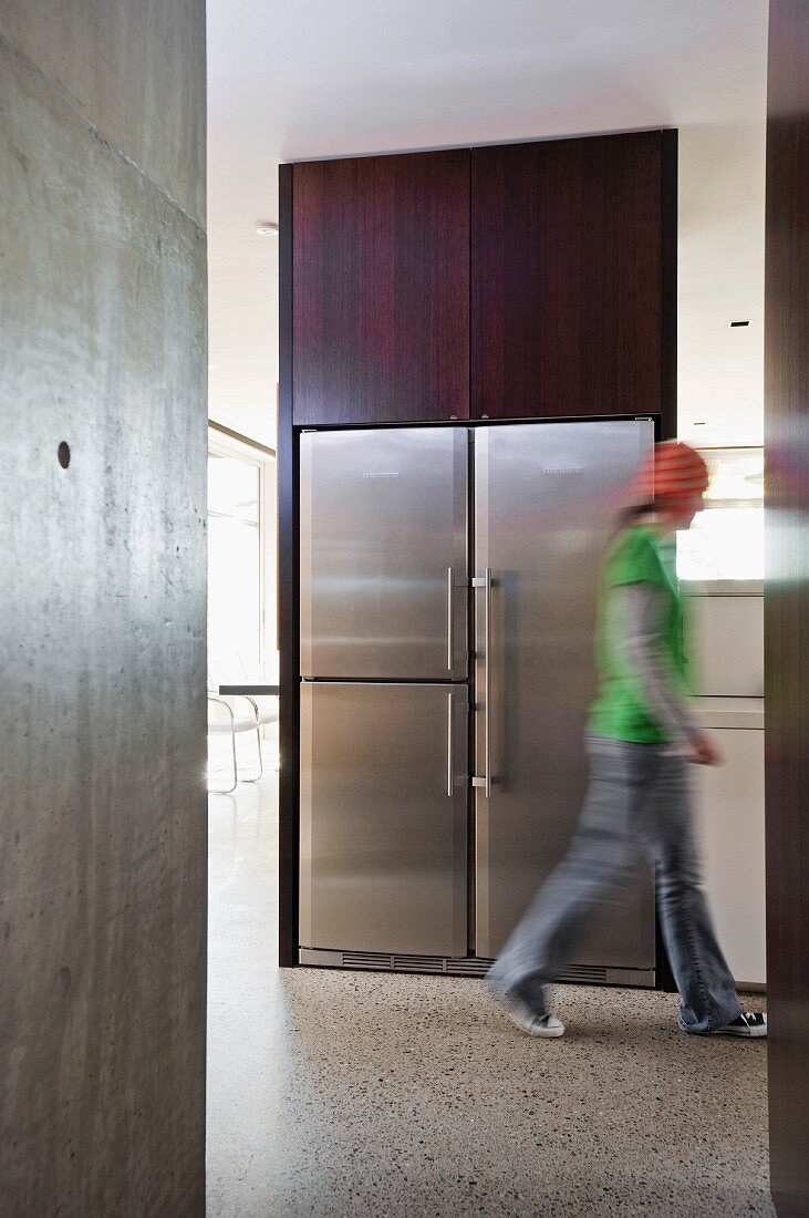 Offener Grundriss in puristischem Wohnhaus mit Sichtbetonwänden - Person läuft vor Einbaukühlschrank mit Edelstahlfronten