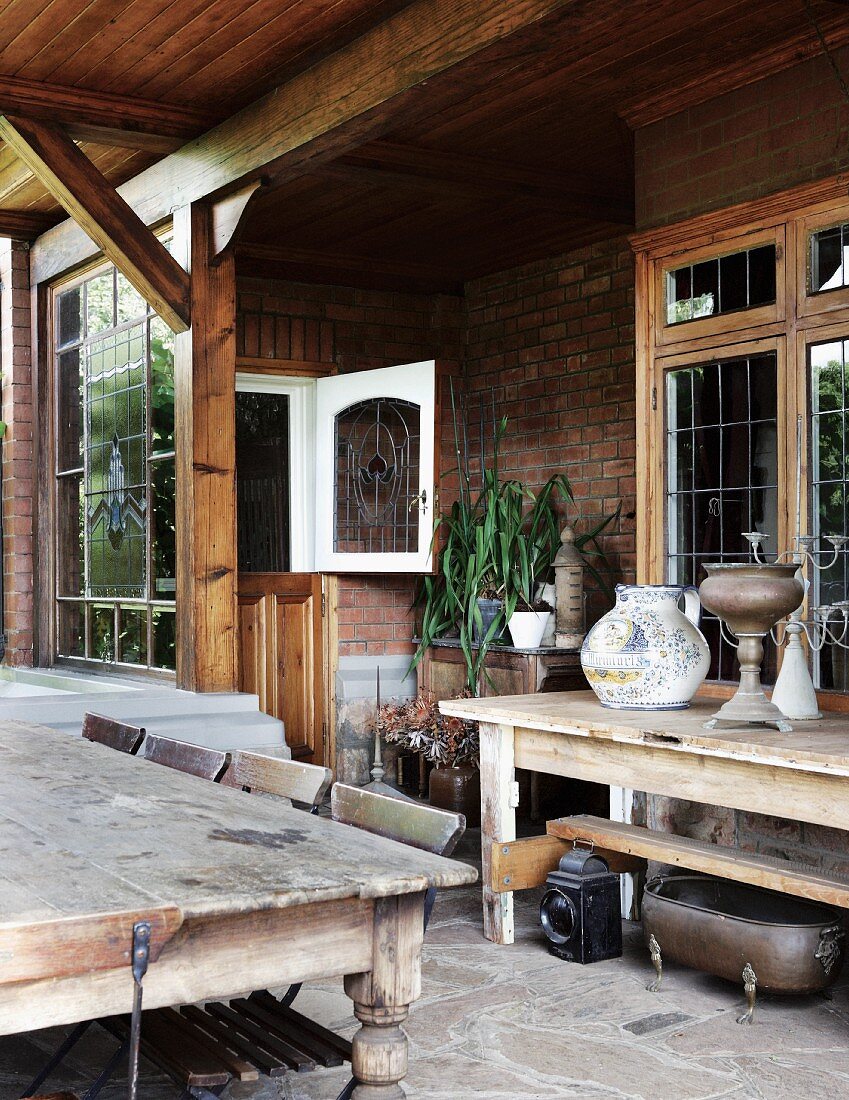 Rustic wooden table with chairs on a covered terrace in front of a country home with a brick facade and Art Nouveau lead glass windows