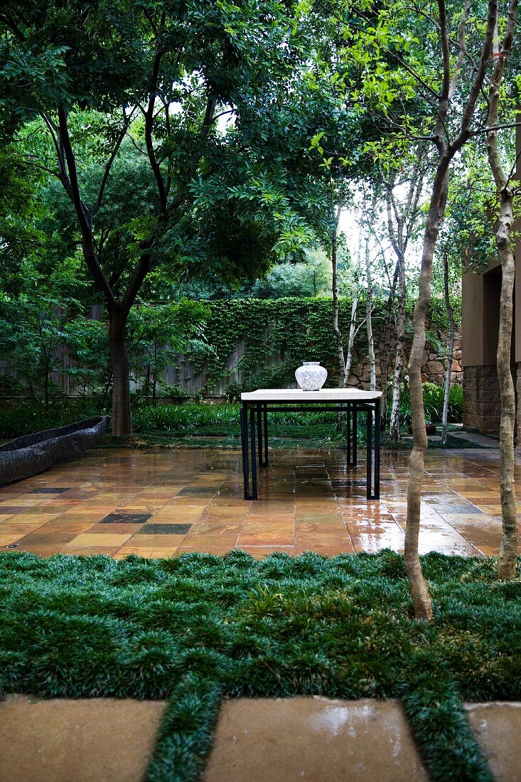 Gartentisch mit Vase auf regennasser, gefliester Terrasse in tropischem Garten