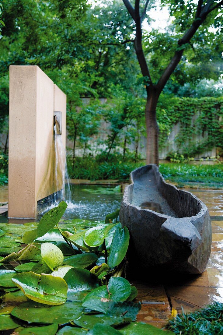 Bootähnliches Objekt aus Stein vor Wasserbecken und Wandscheibe mit Wasserspeier in gestaltetem Garten