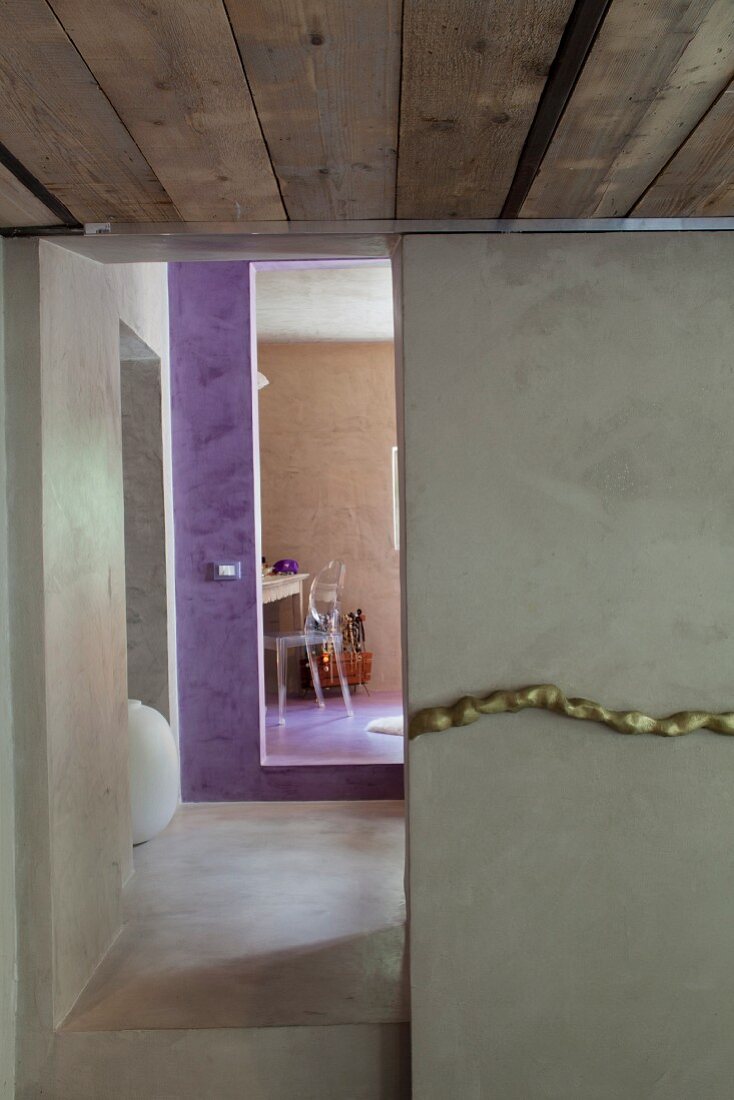 Polierter Beton in Gangflucht mit violett getöntem Badezimmerzugang am Ende; stilisierte Goldkordel als Wanddeko im Vordergrund