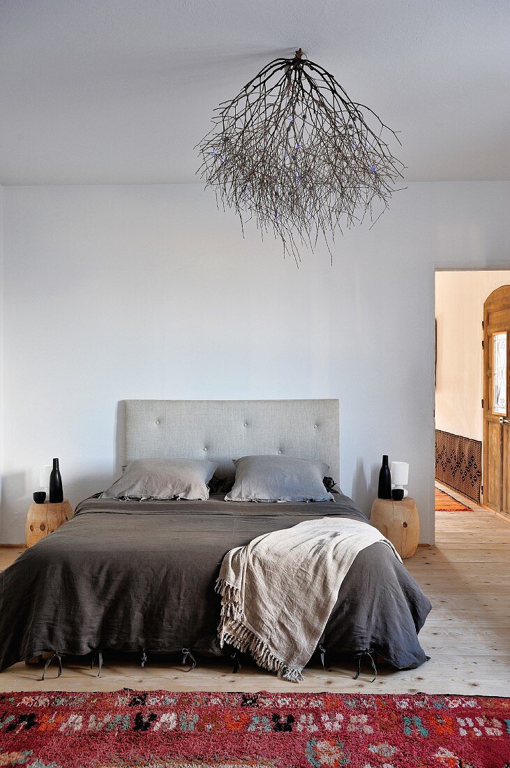 In warmen Grautönen bezogenes französisches Bett, roter Wollteppich und trockener Zweig als naturhafter Lampenschirm an der Decke