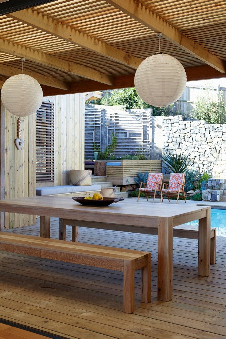 Holztisch mit Bänken und kugelförmige Hängelampen auf einer überdachte Terrasse vor Pool