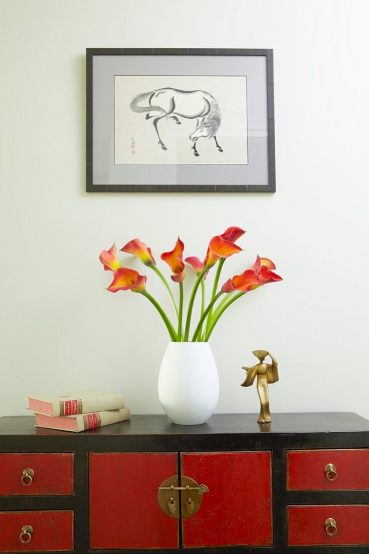 Vase of calla lilies on red and black, Oriental-style sideboard below framed drawing of horse on wall