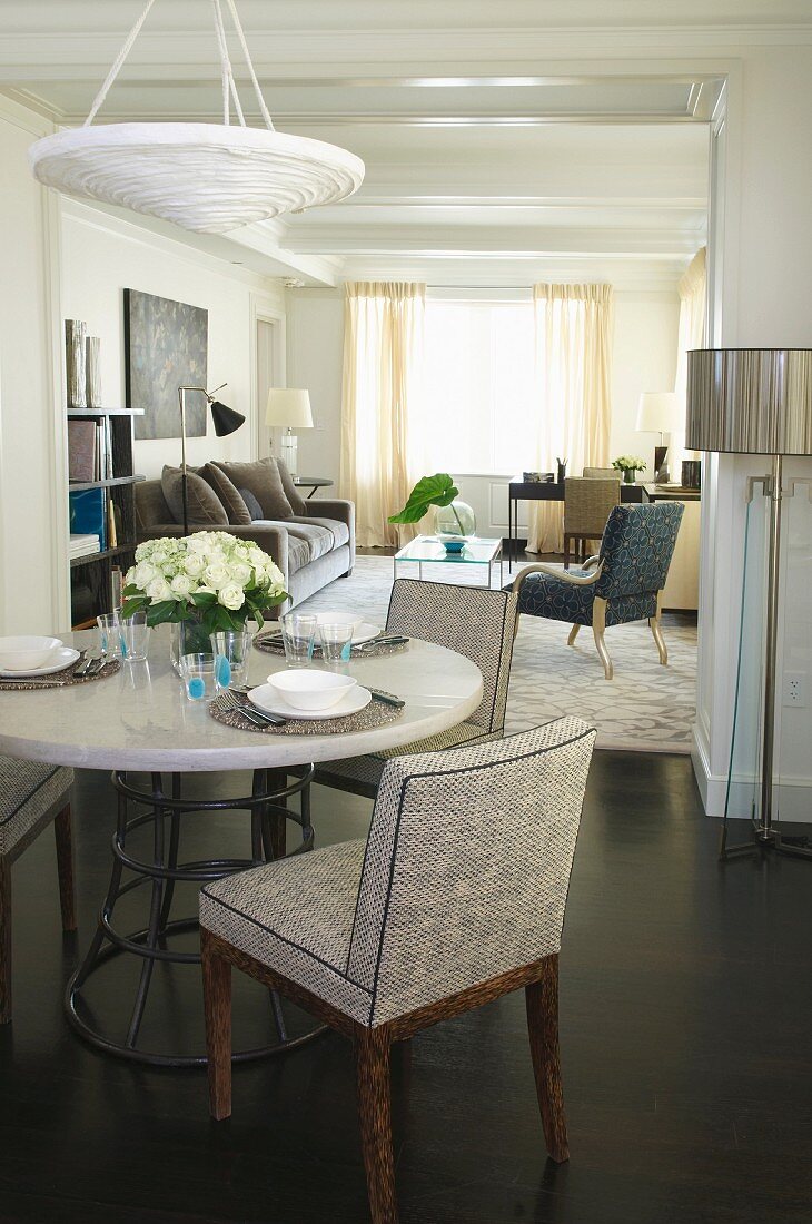 Gedeckter Esstisch und gepolsterte Stühle unter moderner Hängelampe vor breitem Durchgang und Blick ins Wohnzimmer