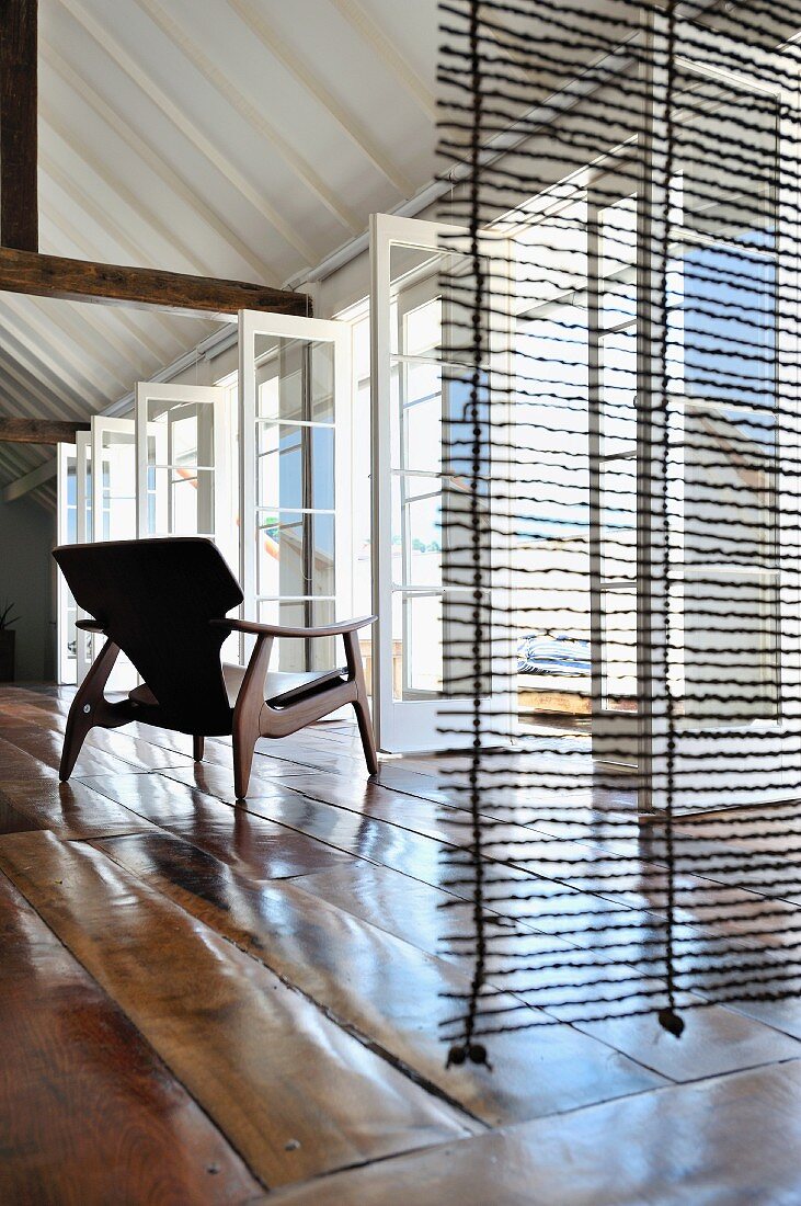Blick durch transparentem Raumteiler auf Sessel mit Holzgestell im Fiftiesstil vor offenstehenden Terrassentüren und rustikaler Dielenboden