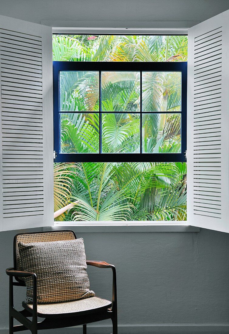 Pillows on an antique chair by a window with open, white interior shutters and a view of palm fronds