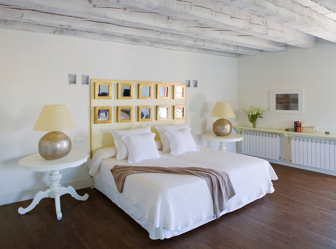 Schlichtes Doppelbett mit gerahmter Spiegelgruppe am Kopfende an Wand zwischen traditionellen Nachttischen im Schlafzimmer mit Holzbalkendecke