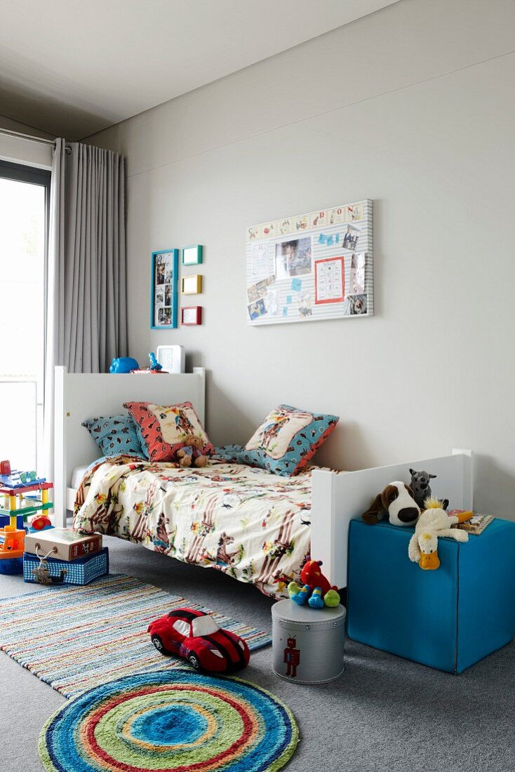 Kinderzimmer mit Spielsachen auf Teppichboden und blauem Sitzwürfel am Bettende vor grauer Wand