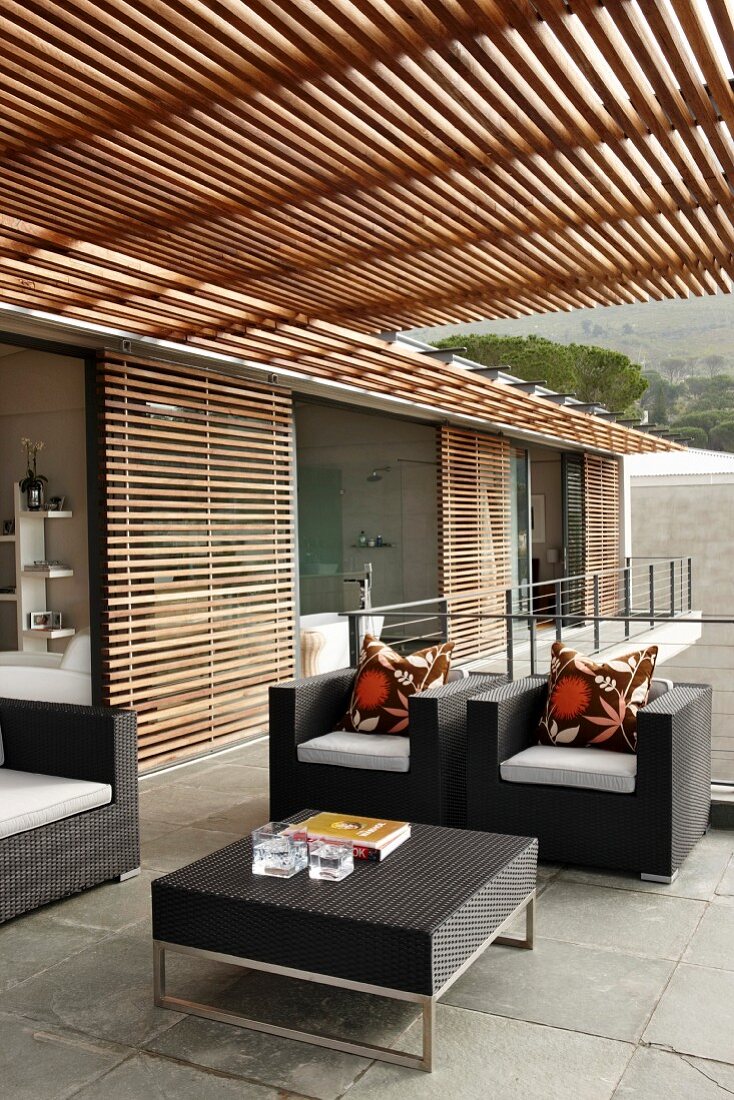 Modernes Wohnhaus mit eleganten schwarzen Outdoormöbeln auf Terrasse unter Pergola aus Holzleisten und Holzjalousien vor der Glasfront