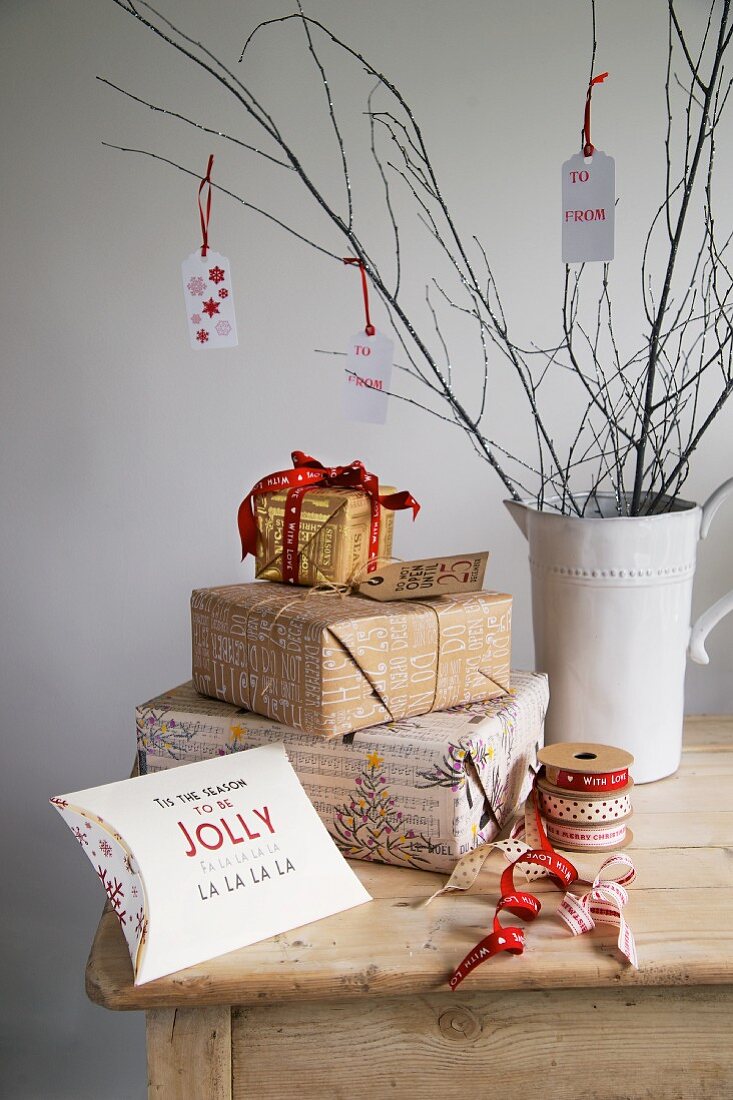 Weihnachtsgeschenke neben Rollen mit Schleifenbändern und weißem Retro-Porzellankrug mit Zweigen & aufgehängten Dekoschildern