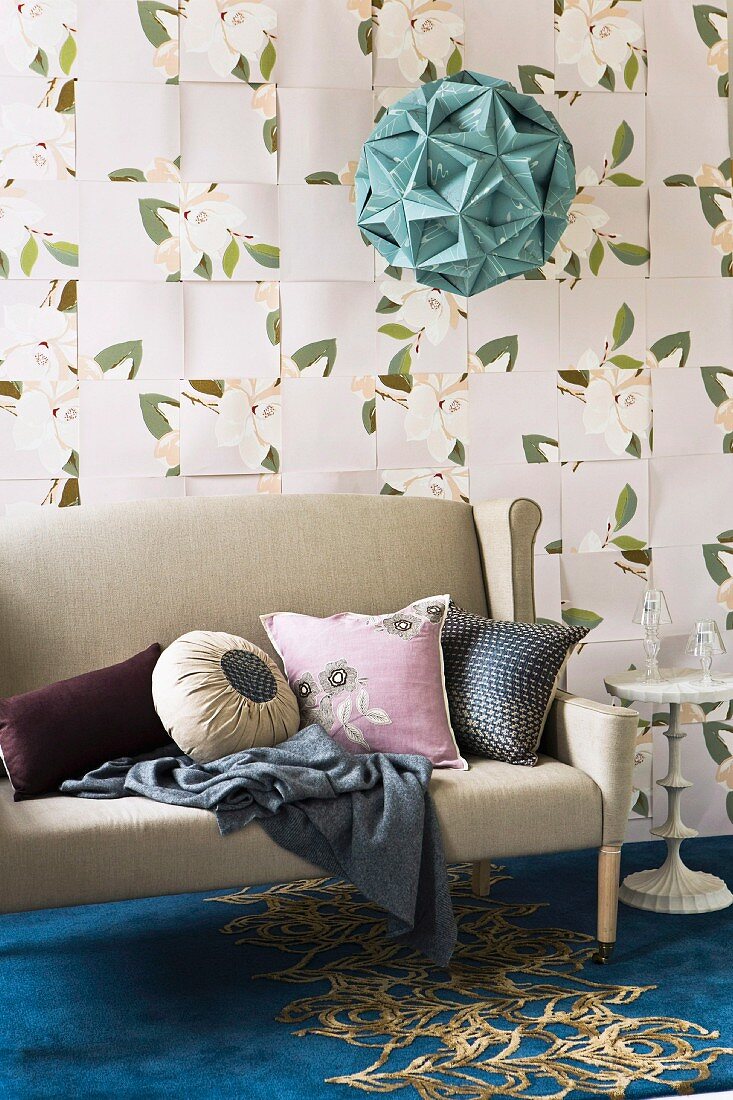 Kissen in verschiedenen Formen und Farben auf hellgrauer Couch auf blauem Teppich mit Golmuster, vor Wand mit Blumenmotiven auf Tapete