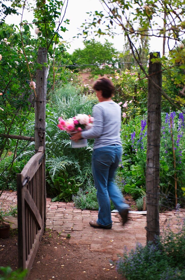 Blick durch offenes Gartentor auf gehende Frau mit Blumenstrauss vor wildem Bauerngarten