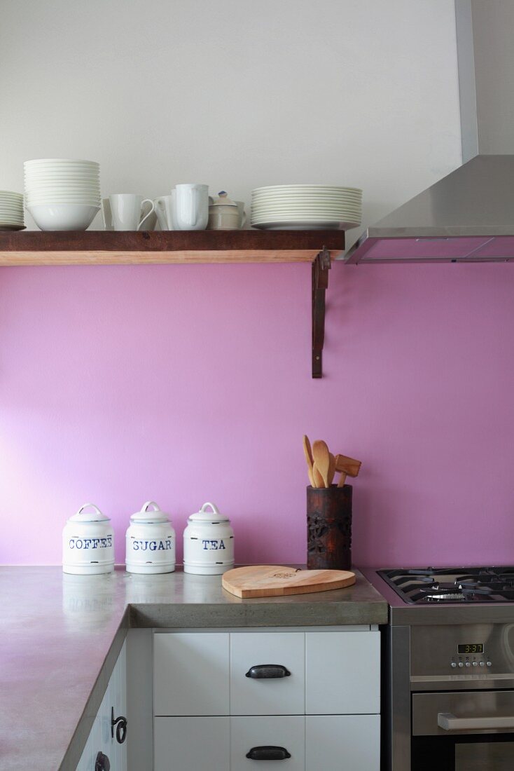 Kitchen counter corner with concrete counter top in front of a violet back splash and white crockery on a wall mounted shelf next to a stainless steel extractor fan