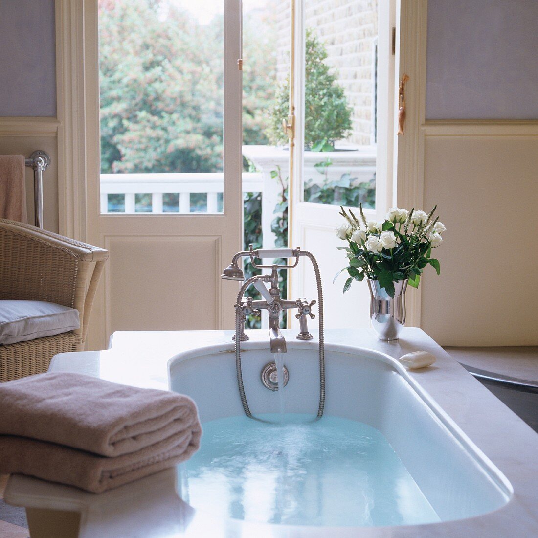 Bathwater in vintage bathtub with vase of white roses on bath surround and view into garden through half-open French doors