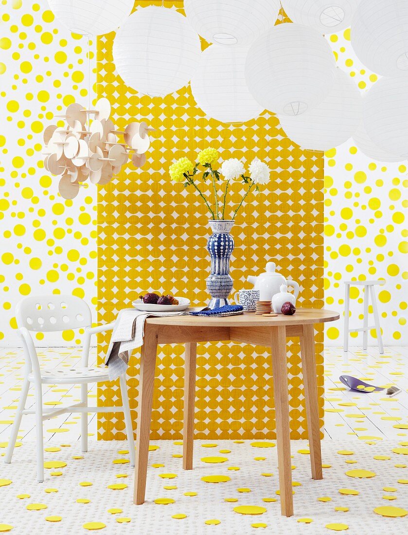 Set wooden table and white plastic chair under round paper lanterns, in front of a room divider with geometric pattern in a room with yellow discs on the floor and wall