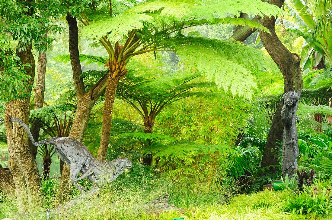 Wildkatzenfigur und Affenskulptur an Baumstamm in dschungelartigem Naturgarten