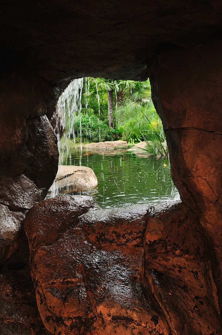 Blick aus der schmalen Öffnung einer Steinhöhle auf eine Teichlandschaft mit Wasserfall