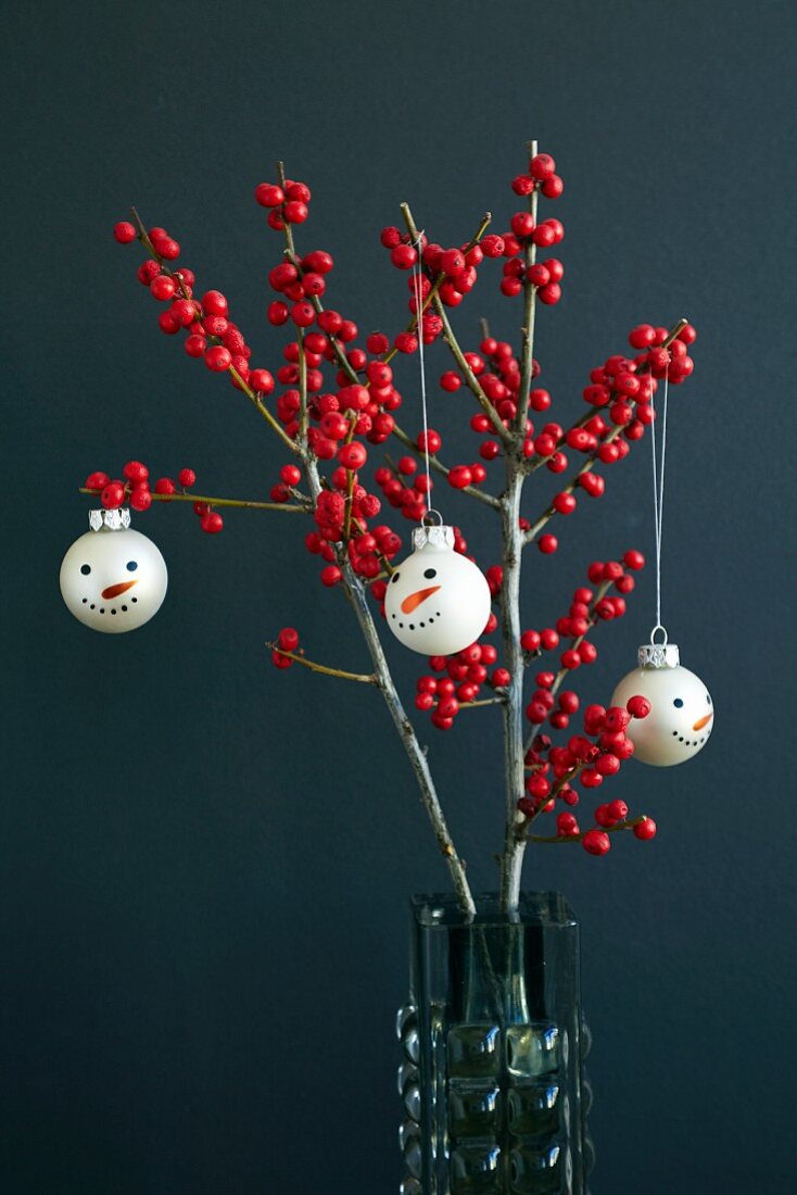Weihnachtsbaumkugeln mit Schneemanngesicht an Stechpalmenzweigen (Ilex) mit roten Beeren
