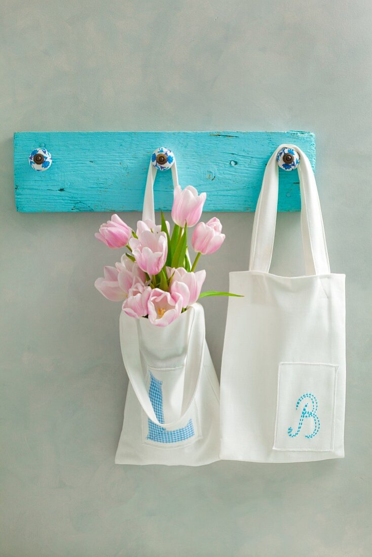 weiße Baumwolltaschen mit den Buchstaben L und B auf einer Hakenleiste, rosa Tulpen in einer Tasche