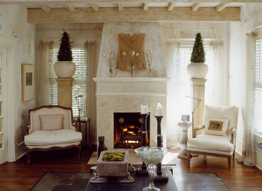 Open fireplace flanked by armchairs in front of decorative bushes in vases on columns against windows in country-style interior