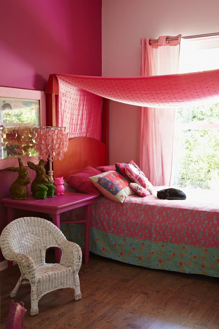 Romantisches Mädchenzimmer in Rottönen mit weißem Rattanstuhl und pinkfarbenem Nachttisch, Bett mit gemusterter Tagesdecke unter Baldachin