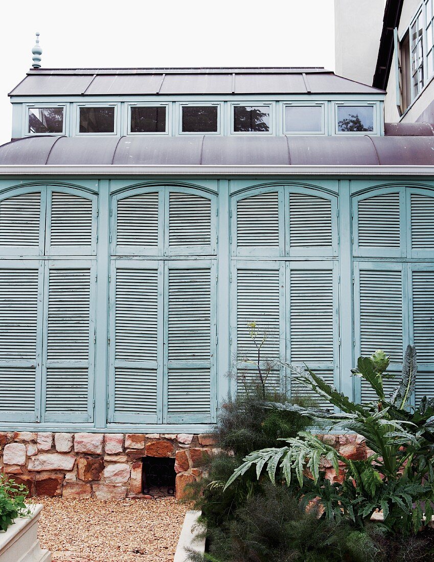Geschlossene, türkisfarbene Fensterläden über Natursteinsockel an einem Vorbau; eingefasste Pflanzbeete zwischen Kieswegen