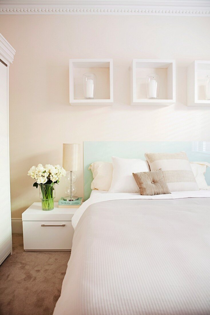 Doppelbett mit Tagesdecke und Kissen in pastelligen Naturfarben vor zart cremefarbener Wand mit weissen Dekorahmen