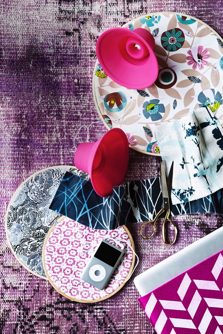 Verschieden gemusterte Stoffe in Stickrahmen und iPod mit pinkfarbenen Silikon-Lautsprechern auf einem Teppich arrangiert